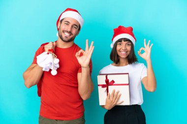 Noel şapkalı genç bir çift mavi arka planda izole edilmiş hediyeler dağıtıyor. Parmaklarıyla iyi bir işaret gösteriyor.