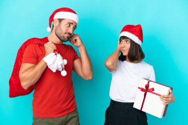 Noel şapkalı genç çift mavi arka planda izole edilmiş hediyeler dağıtırken kafaları kaşınırken kuşkuları oluyor.