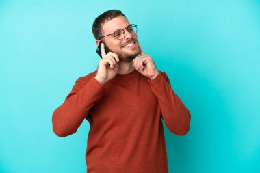 Mavi arka planda izole edilmiş cep telefonu kullanan genç Brezilyalı bir adam yukarı bakarken bir fikir düşünüyor.