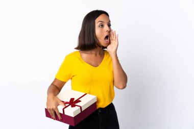 Beyaz arka planda izole edilmiş bir hediye tutan genç Latin kadın ağzı sonuna kadar açık bağırıyor.