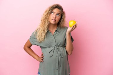 Kıvırcık saçlı kız pembe arka planda izole edilmiş hamile ve elma tutarken hayal kırıklığına uğramış.