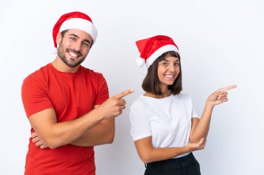 Beyaz arka planda izole edilmiş Noel şapkalı genç çift yan pozisyonu işaret ediyor.