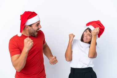 Beyaz arka planda izole edilmiş Noel şapkalı genç çift galibiyet pozisyonunu kutluyor.