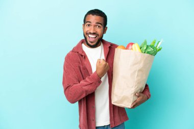 Mavi arka planda izole edilmiş alışveriş torbası tutan genç latin adam zaferi kutluyor.