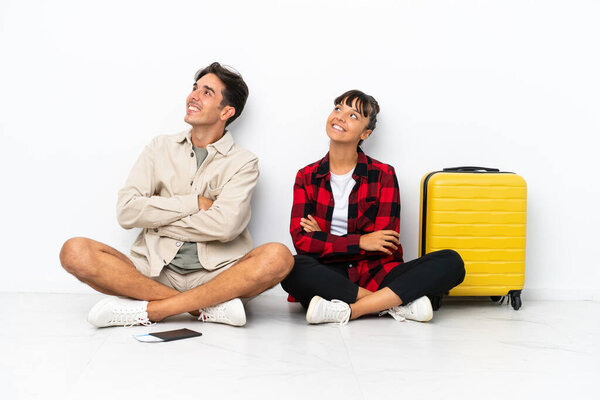 Young mixed race travelers couple sitting on the floor isolated on white background looking up while smiling