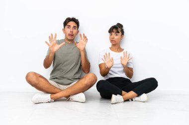 Beyaz arka planda soyutlanmış bir şekilde yerde oturan genç melez çift biraz gergin ve korkmuş bir şekilde öne doğru uzanıyor.