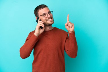 Mavi arka planda izole edilmiş cep telefonu kullanan genç Brezilyalı adam harika bir fikre işaret ediyor.
