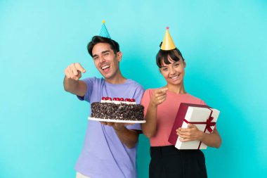 Genç melez bir çift ellerinde doğum günü pastası ve mavi arka planda izole edilmiş bir hediye tutarken gülümserken parmaklarını sana gösteriyor.