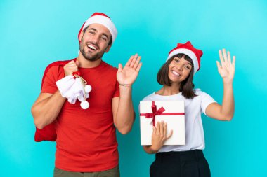 Noel şapkalı genç çift mavi arka planda izole edilmiş hediyeler dağıtıyor mutlu bir ifadeyle selamlıyorlar.