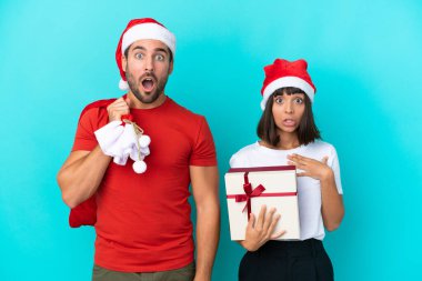 Yılbaşı şapkalı genç bir çift mavi arka planda izole edilmiş hediyeler dağıtıyor. Şaşırmış ve şok olmuş bir yüz ifadesi var.