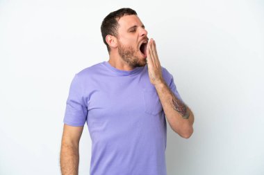 Genç Brezilyalı bir adam beyaz arka planda izole edilmiş esniyor ve geniş ağzını eliyle kapatıyor.