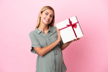 Genç Uruguaylı kadın mavi arka planda izole edilmiş hamile ve elinde bir hediye var.