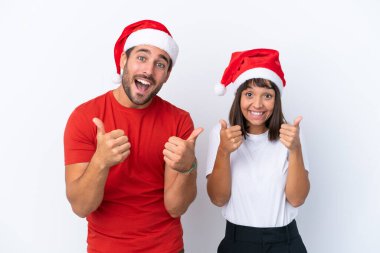 Beyaz arka planda izole edilmiş Noel şapkalı genç bir çift iki eliyle baş parmak işareti yapıyor ve gülümsüyor.