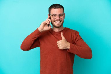 Mavi arka planda izole edilmiş cep telefonu kullanan genç Brezilyalı adam işaret veriyor.