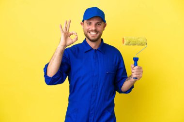 Ressam Brezilyalı adam sarı arka planda izole edilmiş, tamam işareti parmaklarıyla gösteriyor.