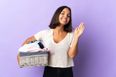 Mor arka planda izole edilmiş çamaşır sepetini tutan genç Latin kadın mutlu bir ifadeyle selamlıyor.