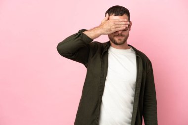 Genç Brezilyalı bir adam pembe arka planda izole edilmiş, gözlerini elleriyle kapatıyor. Bir şey görmek istemiyorum.