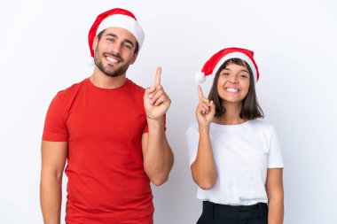 Beyaz arka planda izole edilmiş Noel şapkalı genç çift en iyinin işareti olarak parmağını kaldırıyor.