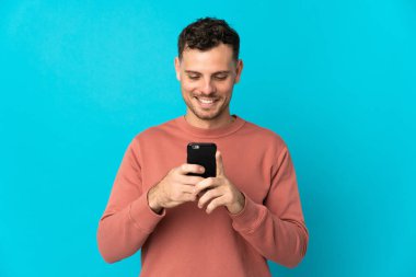 Mavi arka planda izole edilmiş genç beyaz adam cep telefonuyla mesaj ya da e-posta gönderiyor.