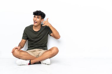 Genç Arjantinli bir adam yerde oturmuş telefon jesti yapıyor. Bana geri dönüş işareti ver.