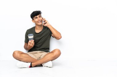 Genç Arjantinli bir adam elinde kahve ve cep telefonu ile yerde oturuyor.