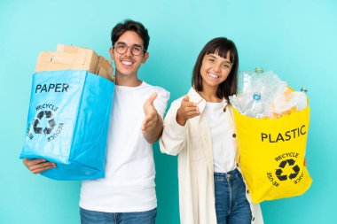 Mavi arka planda izole edilmiş plastik ve kâğıt dolu bir çanta tutan genç çift iyi bir anlaşma için el sıkışıyor.