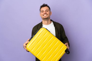 Beyaz arka planda izole edilmiş beyaz tenli yakışıklı bir genç seyahat valiziyle geziyor.