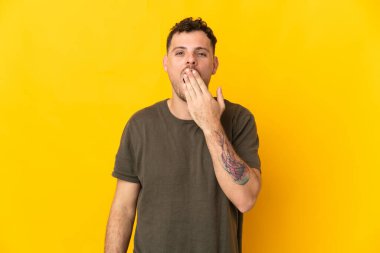 Beyaz tenli yakışıklı adam sarı arka planda izole edilmiş esniyor ve ağzını eliyle kapatıyor.