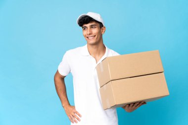 Teslimatçı Arjantinli adam mavi arka planda izole edilmiş. Kolları kalçasında ve gülümsüyor.