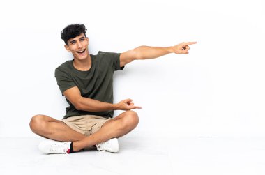 Genç Arjantinli bir adam yerde oturuyor. Parmakla göstererek bir ürün sunuyor.