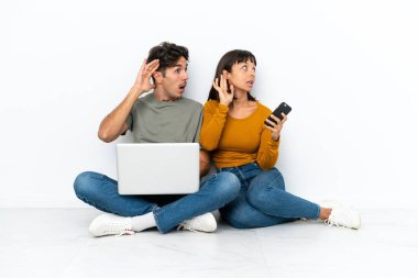 Dizüstü bilgisayarı ve cep telefonu olan genç bir çift yerde oturup bir şeyler dinliyorlar.