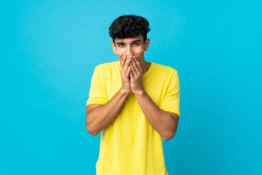 Arjantinli genç adam arka planda izole edilmiş mutlu ve gülümseyen elleriyle ağzını kapatıyor.