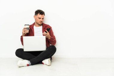 Genç, yakışıklı, beyaz bir adam dizüstü bilgisayarıyla yerde oturuyor. Elinde kahve ve cep telefonu var.
