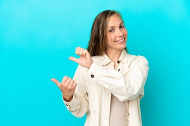 Mavi arka planda izole edilmiş genç beyaz kadın bir ürünü sunmak için kenarı işaret ediyor.