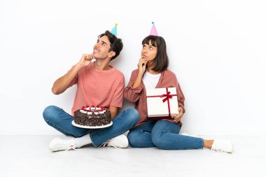 Genç melez çift, beyaz arka planda izole bir şekilde yerde oturmuş doğum gününü kutlarken kafalarını kaldırıp bir fikir üretiyor.