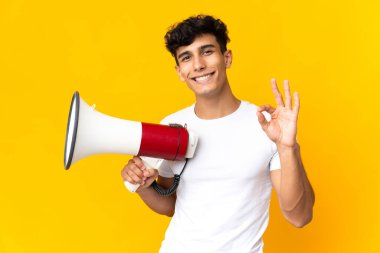 Sarı arka planda izole edilmiş genç Arjantinli bir adam elinde bir megafonla tamam işaretini parmaklarıyla gösteriyor.
