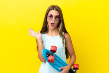 Küçük beyaz kaykaycı kız sarı arka planda izole edilmiş ve sürpriz jest yapıyor.