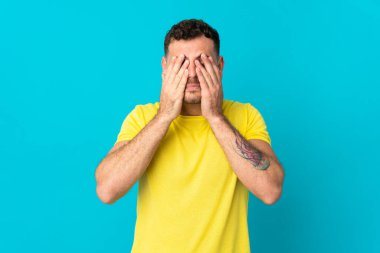 Beyaz tenli yakışıklı adam mavi arka planda izole edilmiş gözleri elleriyle kapatıyor.