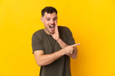 Sarı arka planda izole edilmiş beyaz, yakışıklı genç bir adam bir ürün sunmak ve bir şeyler fısıldamak için kenarı işaret ediyor.
