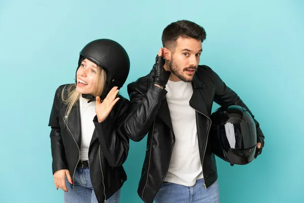 Couple with motorcycle helmet over isolated blue background listening to something by putting hand on the ear