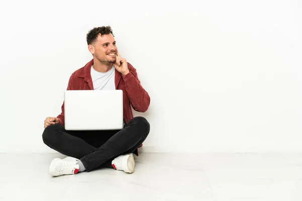 年轻英俊的高加索男人坐在地板上 一边看一边用笔记本电脑思考着一个想法 — 图库照片