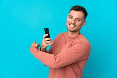 Genç, beyaz, yakışıklı bir adam mavi arka planda izole edilmiş, cep telefonu kullanıyor ve onu işaret ediyor.