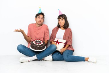 Genç melez çift, beyaz arka planda izole bir şekilde yerde oturmuş doğum gününü kutluyorlar. Omuzlarını kaldırırken şüphe duyuyorlar.