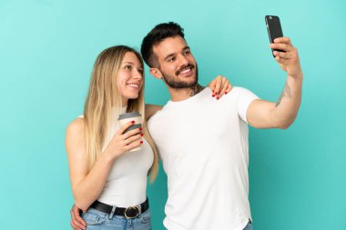 İzole edilmiş mavi arka planda genç bir çift cep telefonuyla selfie çekiyor.