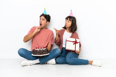 Genç melez çift, beyaz arka planda izole bir şekilde yerde oturmuş şaşırmış bir yüzle parmak göstererek doğum gününü kutluyor.