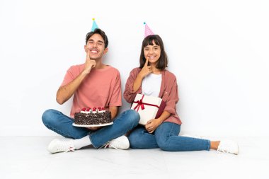 Genç melez çift, beyaz arka planda izole edilmiş bir şekilde yerde oturmuş mutlu ve hoş bir ifadeyle gülümsüyor.