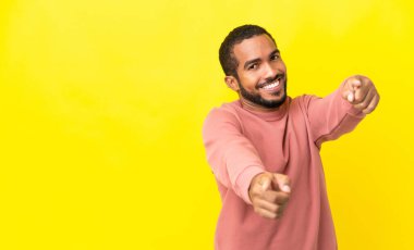 Sarı arka planda izole edilmiş genç latin adam mutlu bir ifadeyle ön tarafı işaret ediyor.