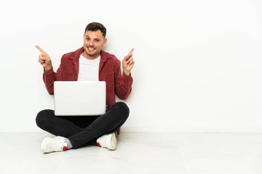 Genç, yakışıklı, beyaz bir adam dizüstü bilgisayarla yerde oturuyor ve parmak uçlarını işaret ediyor.
