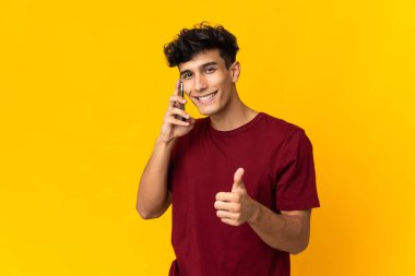 Arjantinli genç adam sarı arka planda izole edilmiş. Cep telefonuyla konuşurken baş parmağını kaldırıyor.