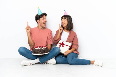 Genç melez çift, beyaz arka planda izole bir şekilde yerde oturmuş doğum gününü kutluyorlar. Çözümün farkına varmak için parmağını kaldırıyorlar.